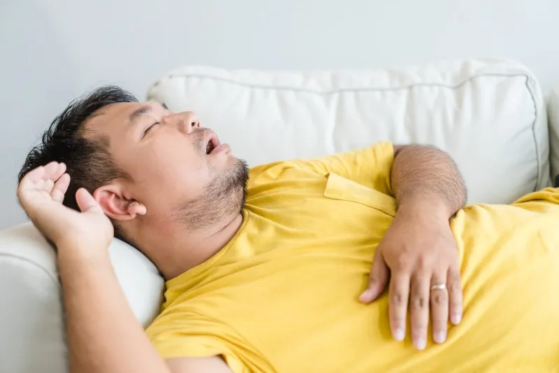 An Asian man snoring while sleeping on a couch